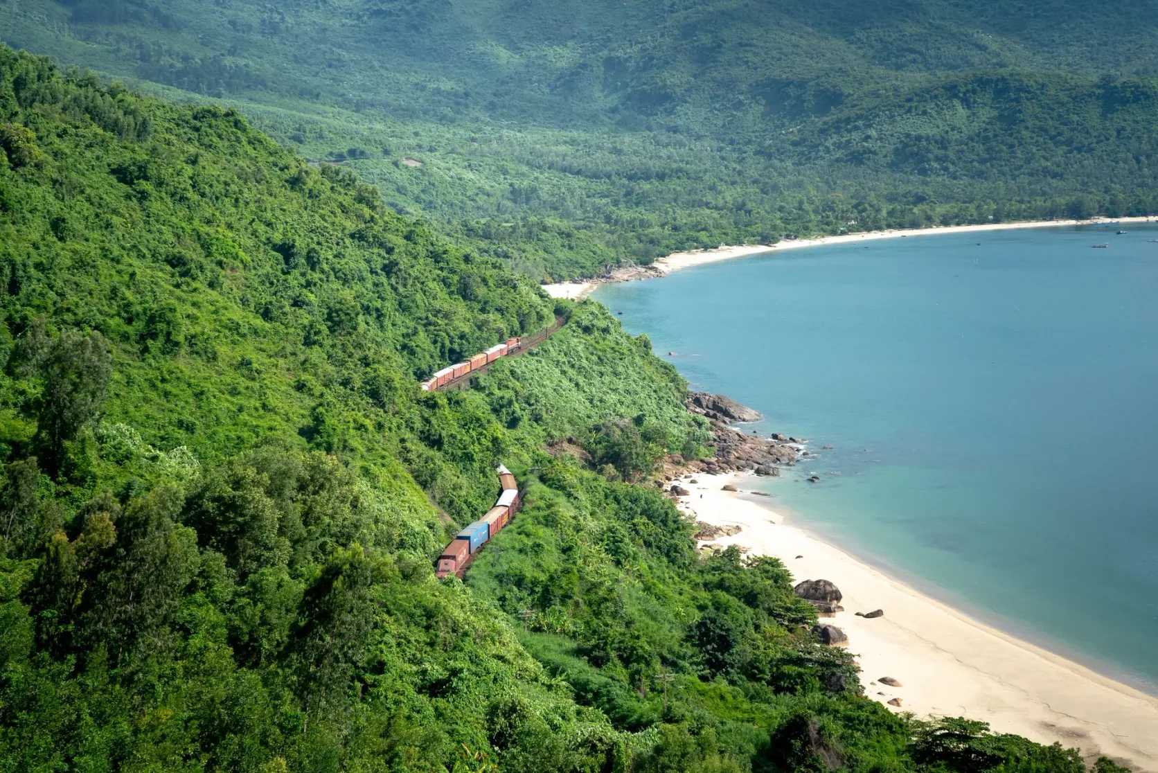 Ein Zug fährt auf der Küstenstrecke zwischen grünen Hügeln und dem Meer.